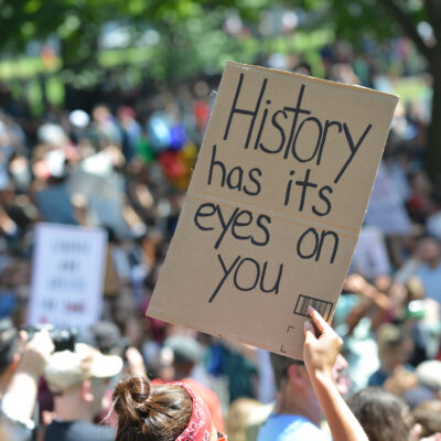 Demonstrasjon hvor en person holder opp en plakat hvor det står: History has it's eyes on you.