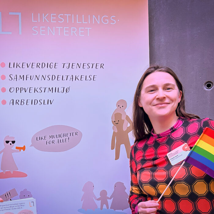Frøydis Sund står foran en rollup med Likestillingssenterets logo. Hun smiler og holder et pride-flagg.